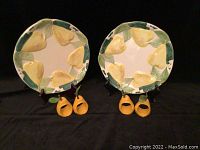 Two ceramic salad plates with hand-painted yellow pear and green leaf design around the rim, displayed with four pear-shaped napkin rings in front of each plate on black background.