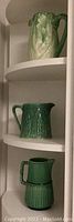 Three green antique French jugs displayed on a white, curved shelf, showing the different shapes and detailed textures of each jug.