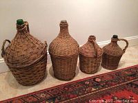 Four wicker-wrapped vintage demijohn bottles of various sizes and shapes, set on carpeted floor next to wall and rug.