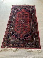 Full view of the reddish Belgian wool rug laid out on carpet. Shows the entire rug and fringe edges, with a central geometric design in various colors.