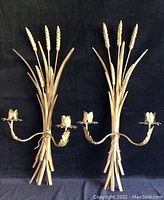 Photo showing two brass wheat wall mounted candle holders against a black background. Both holders have two candle arms with flower-shaped candle cups and wheat stalk details.