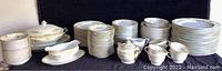 Full set displayed showing stacks of plates, bowls, cups, and serving pieces arranged on a black surface with white and floral gold trimmed china.