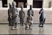 Photo of 5 Chinese clay soldiers and 1 horse figure, arranged on a reflective surface with a neutral background.