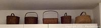 Five antique woven baskets displayed on a white shelf against a wall. The baskets vary in shape from rectangular to oval to rounded, each with a handle on top. Colors are various shades of brown, natural wicker.