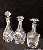 Photo showing three clear crystal decanters each with a distinct shape and silver nameplate; one stopper is cracked.