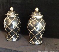 Photo showing two tall black jars decorated with gold rope garland pattern forming diamond shapes and topped with matching lids with gold finials.