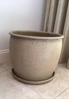 Front view of large tan off-white clay plant pot with water tray on tiled floor near curtain.