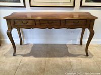 Full front view of the oak side table with three drawers and curved legs on tiled floor.