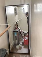 Full frontal view of large rectangular framed mirror reflecting a narrow room with various objects including a water heater and some tools.