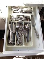 Drawer with stainless steel flatware including knives, forks, spoons, and soup ladle organized in white tray.