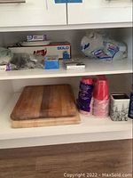 Shelf showing wooden cutting boards, disposable cups, and packaged paper plates and plastic utensils