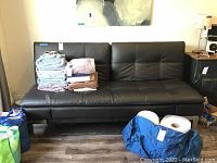 Full front view of the black futon couch with some folded towels and household items on top and nearby.