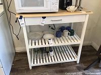White wooden kitchen work surface with light wood top, single drawer with silver handle, and two slatted shelves. Various kitchenware including plates, bowls, mugs, and glasses are placed on and under shelves.