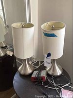 Two metal base lamps with white shades on a round black table with a small red and black alarm clock and cords.