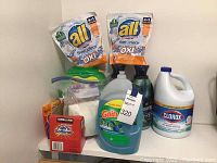 Photo showing multiple brand name laundry cleaning products including All Free Clear Oxi detergent refill bags, Gain liquid detergent, Clorox bleach, Downy fabric softener, Kirkland fabric softener powder and scent boost beads containers.