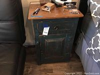 Wooden shabby chic nightstand with blue and brown paint, single drawer with metal ring pull, cabinet door with knob, next to a bed and chair on wood flooring.