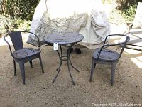 Full set outdoor metal table with two matching metal chairs, set on gravel surface with covered object in background