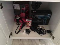 View inside cabinet showing multiple hair dryers including Revlon and another brand in boxes, several attachments and a Norelco men's grooming product.