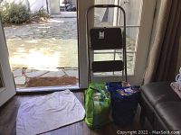 Photo shows a black 2-step folding step stool near a glass door, white cleaning rag laid out on the floor, and two reusable bags (green and blue) filled with assorted cleaning cloths/rags.