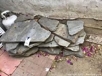 Stack of natural stone paver slabs arranged by size against an exterior wall, showing rough texture and gray coloration.