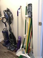 Full view of cleaning tools in corner: two upright vacuums (Hoover Optima and Bissell Rocket), multiple brooms, mops, white dustpans, mop heads with cloths