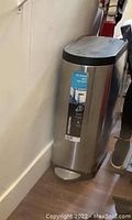 Photo showing the trash can standing upright against a wall, foot pedal visible at the base, stainless steel body with a black lid, label sticker intact on front.