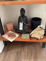 Photo showing the 15-inch tall dark wood Buddha statue seated with wooden bead necklace, black vase to right, patterned boxes, and cloth items on a wooden shelf.