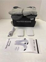 Slider with one pair of gray heated socks, two rectangular white battery packs, a USB cable, and instruction manual on white background