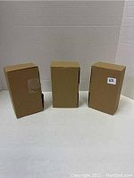 Three sealed simple brown cardboard boxes standing upright on a white surface against a white backdrop. One box has a small label with '1/2' written on it.