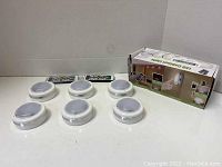 Photo showing six white LED cabinet puck lights, two rectangular remote controls, and original product box grouped on a white surface