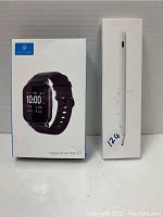 Photo showing Haylou Smart Watch 2 box to the left with image of watch and black band, and white stylus pen box to the right, both boxes upright on a white surface.