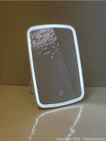 Front view of the rectangular makeup mirror lit with white LED light around edges, showing the touchscreen power button on the lower center of the mirror surface.