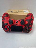 Front view of a red camouflage wireless PS4 controller with all buttons, analog sticks, and touchpad visible, in front of a box.