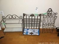 Three iron shelving/storage pieces with decorative ironwork and glass shelves shown on wooden floor against white wall.