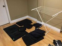 Photo of six black bathroom mats of assorted sizes along with folded black towels and a white collapsible drying rack positioned on floor in corner.
