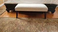 Front view of the beige upholstered bench with nailhead trim and dark wood legs placed in front of a bed on a wooden floor with a patterned rug.