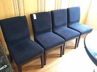 Four faux leather dining chairs with dark wood legs, covered with fitted black fabric seat and back covers. Chairs aligned in row, placed on hardwood floor next to wood-paneled wall.