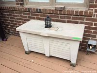 View of outdoor white plastic deck box placed against brick wall on deck, showing front and top with crack on lid.