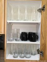 Cabinet shelf with 4 tall beer glasses on top shelf, 3 plastic tumblers and 4 black mugs on middle shelf, 6 clear tumblers on bottom shelf