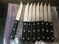 Set of eight Henckels steak knives laid out side-by-side, showing black handles with three rivets and a mix of serrated and straight blades.