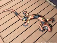 Full view of hammered metal gecko wall decor, showing the whole lizard with curled tail and multi-color hammered metal finish on a wooden floor.