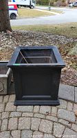 Black square resin outdoor planter shown from front angle on paved outdoor area with natural surroundings.