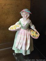 Front view of Royal Doulton Daffy-Down-Dilly figurine showing woman holding two baskets of yellow daffodils, dressed in pastel green and pink with bonnet.