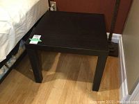 Photo of one black MDF side table with removable legs, placed on wood floor next to bed.