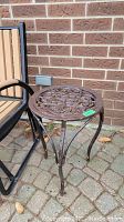 Full view of round metal outdoor side table showing decorative scrollwork tabletop and curved legs against brick wall background.