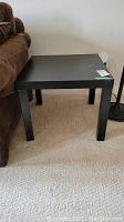 Black square side table placed next to a brown couch and a black floor lamp, showing flat top and four legs with no visible defects