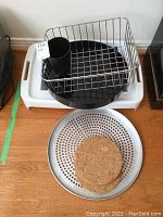 White plastic serving tray on bottom with black round plastic tray and metal wire dish drainer on top including black plastic cutlery holder. Perforated metal tray on floor with three cork trivets stacked inside.