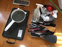 Overall view of kitchen utensils grouped together with spatulas, ladles, whisks, scoops, scissors, and other small tools. Two black rectangular baking pans stacked side-by-side with a metal mesh strainer resting on one.