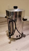 Full view of the stainless steel coffee urn with lid on showing side handles, spout, and power cord coiled on side.