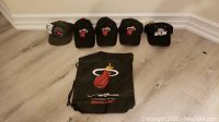 Five basketball hats displayed in a row with a Miami Heat drawstring bag in front